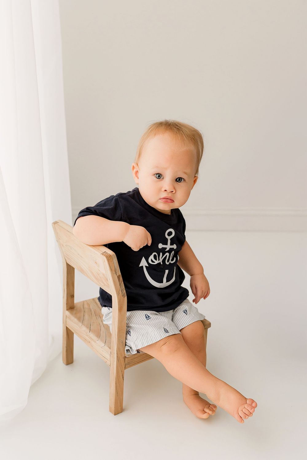 Baby boy in navy anchor first birthday shirt sitting on wooden bench in white studio during milestone session at Summerville SC