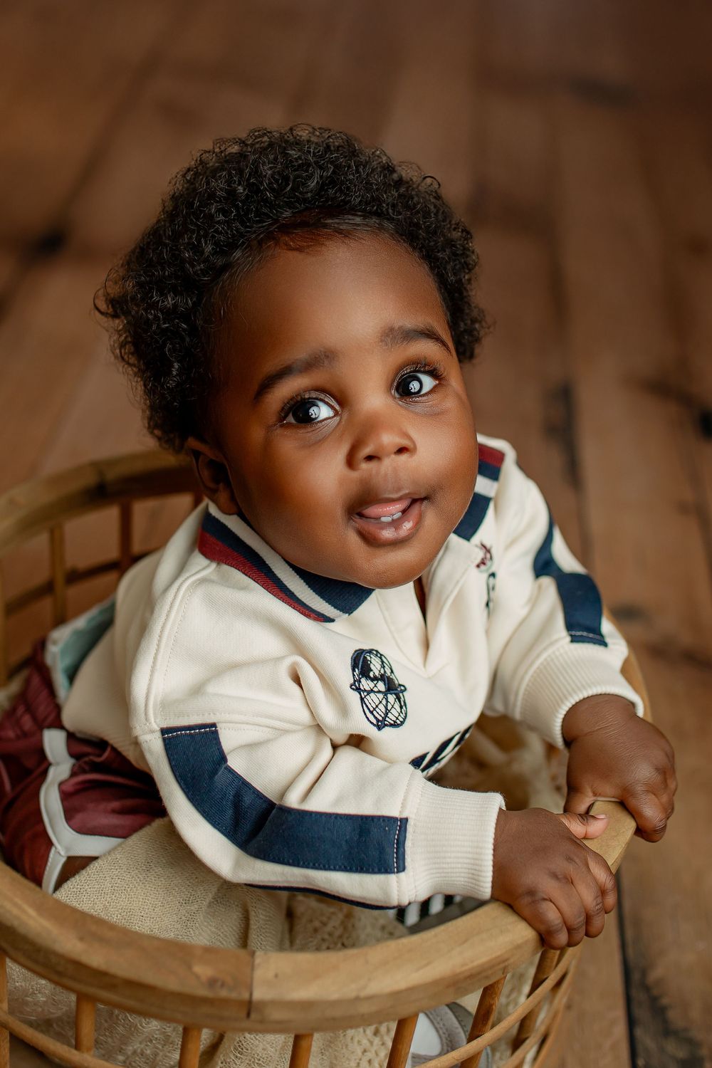 Baby boy in cream and navy varsity sweater looking up from wicker basket on dark wood floor during sitter portrait in Summerville SC
