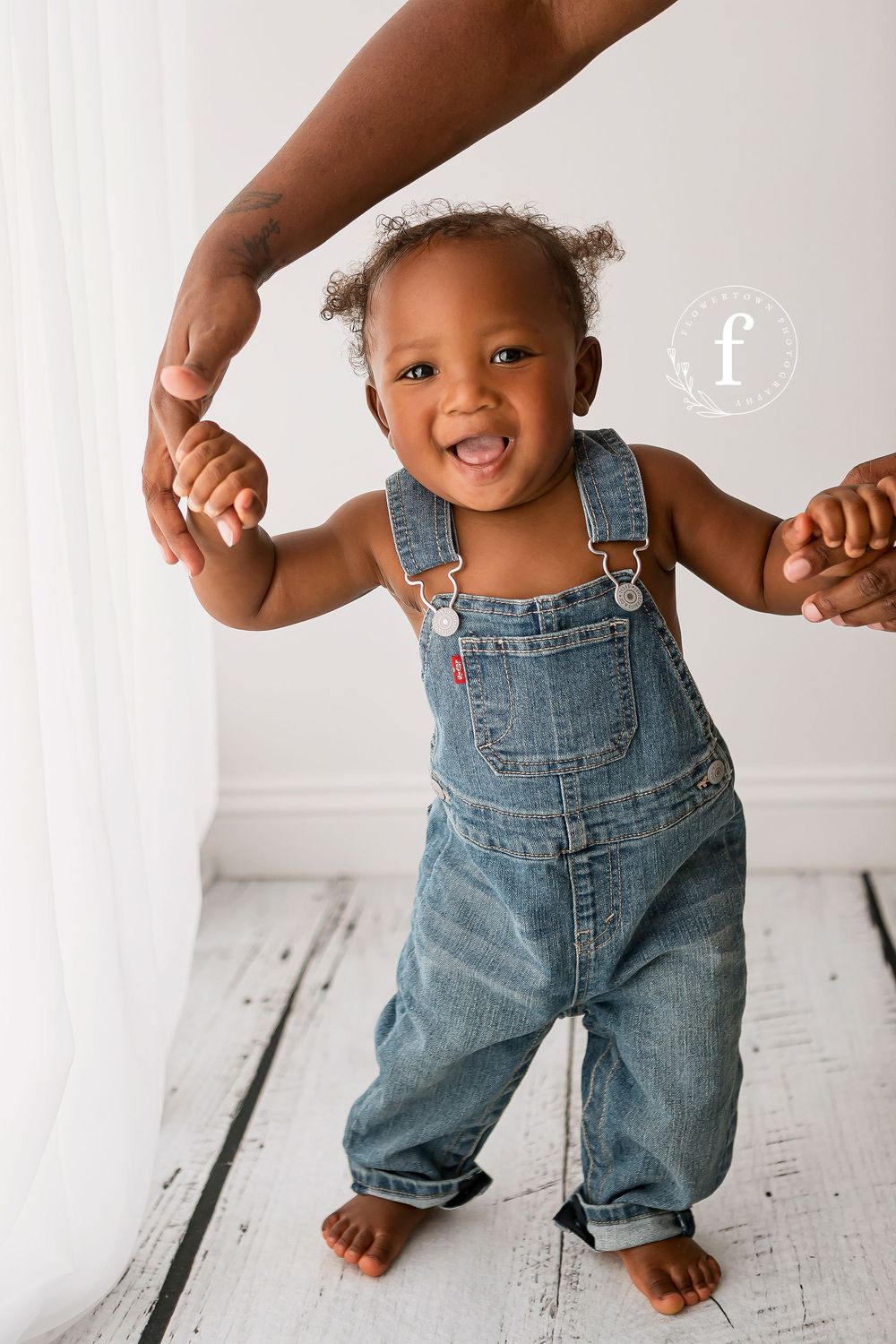 Smiling toddler in denim overalls taking first steps holding parents hands on white wood floor during milestone session in Summerville SC