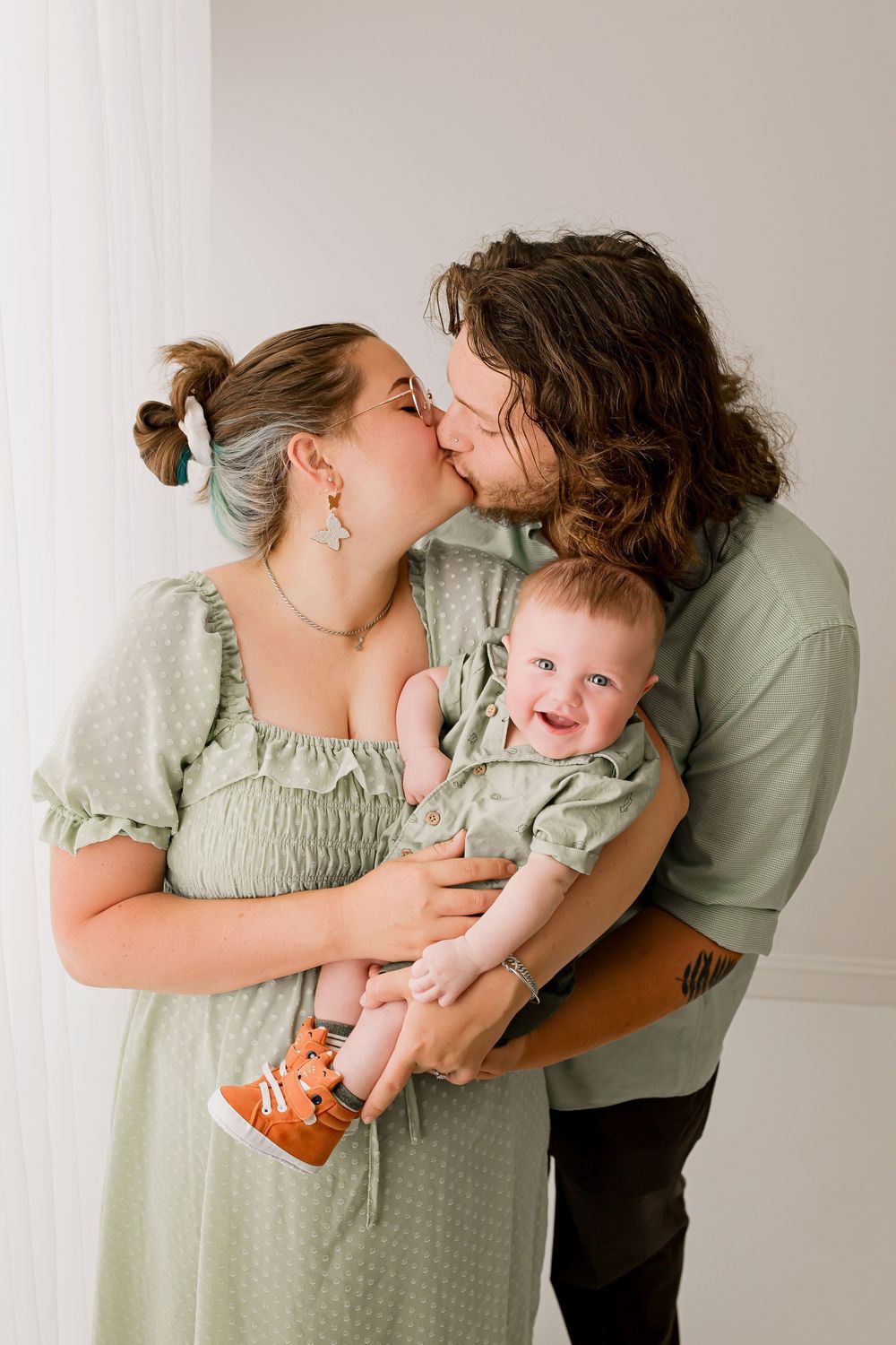 Parents in sage green kissing while holding smiling baby during family milestone session at Flowertown Photography in Summerville SC
