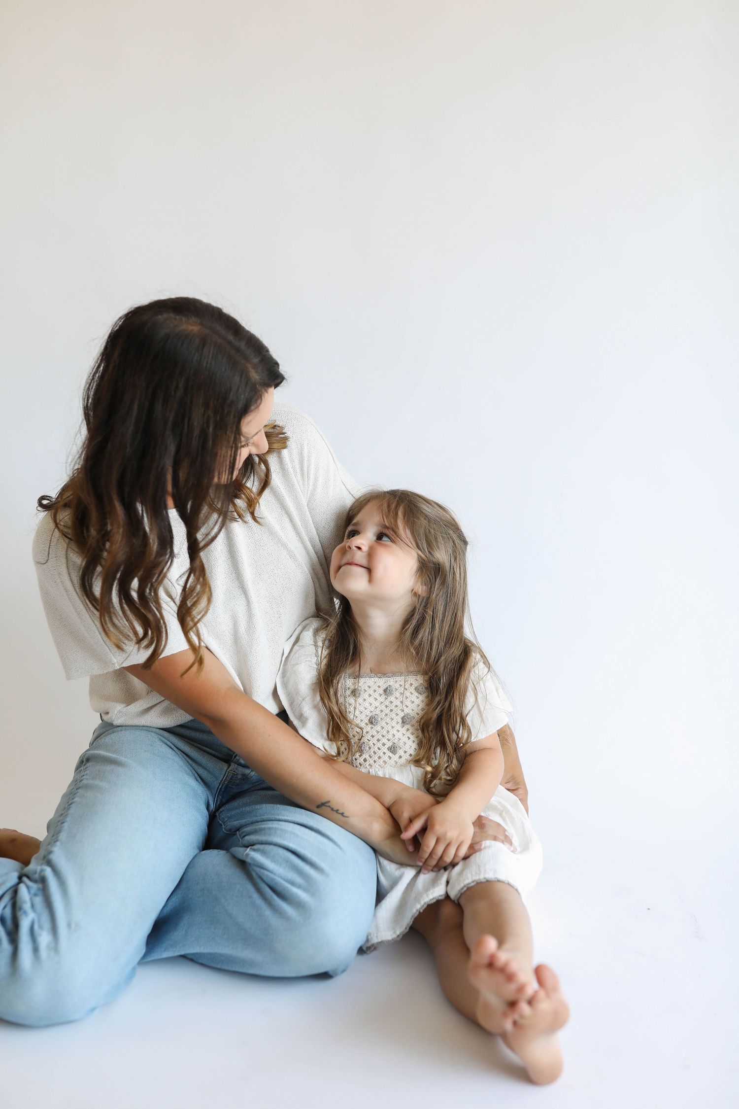 Brunette mother in white tee and jeans sitting on floor smiling at young daughter in white dress.