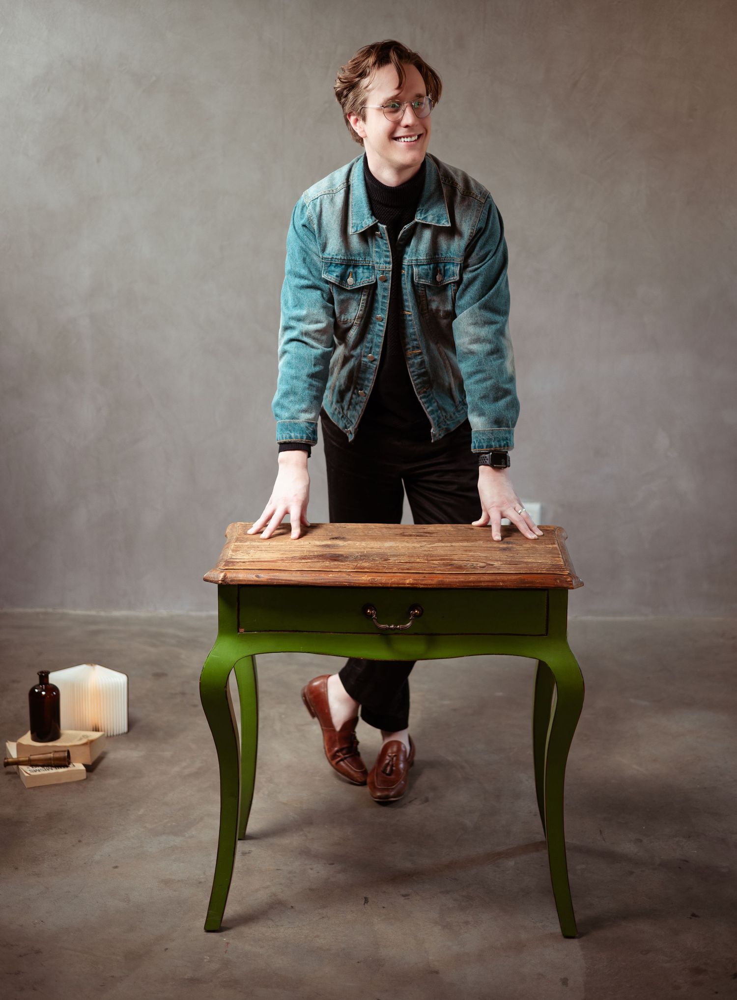 Person standing upside down with hands on wooden table with green painted legs wearing denim jacket against gray background.