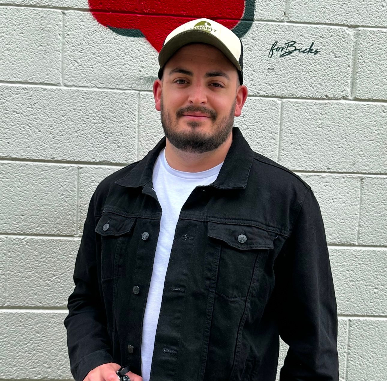 Person in dark jacket and red baseball cap stands against white brick wall while holding up finger in casual pose.