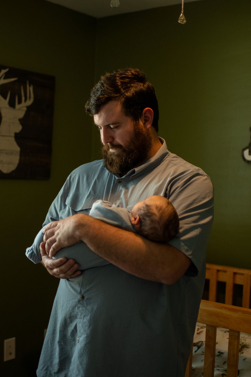 Bearded father tenderly holds newborn baby in nursery with green walls and deer decor.