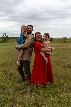 A family embraces together in a grassy field on an overcast day, wearing red dresses and earth-toned clothing.