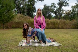 Two young women pose on a striped blanket in a grassy park, one in pink sweater and jeans, surrounded by trees.