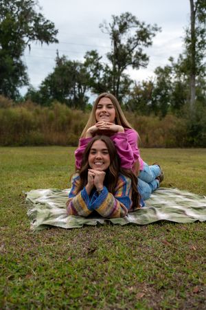 Two smiling girls lying on a blanket outdoors, posing with chins resting on hands in a grassy park setting.