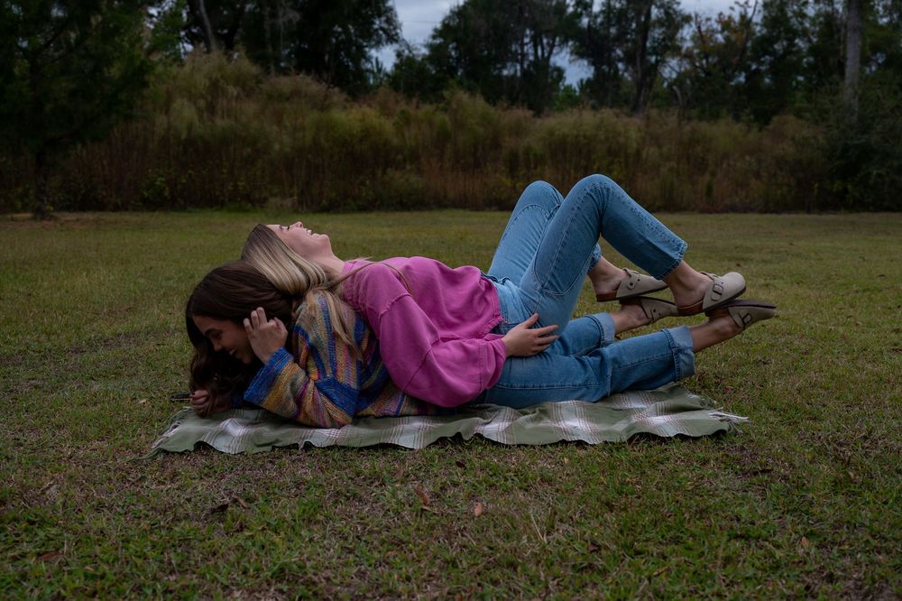 Two women lying on a blanket in a grassy field, cuddling together outdoors.