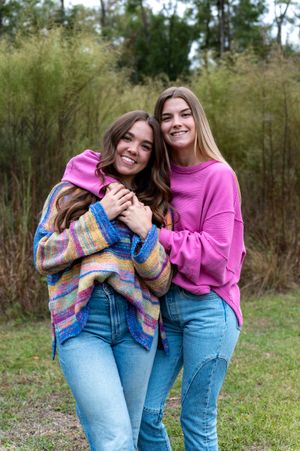 Two smiling young women in pink tops and jeans pose together outdoors with green trees in the background.