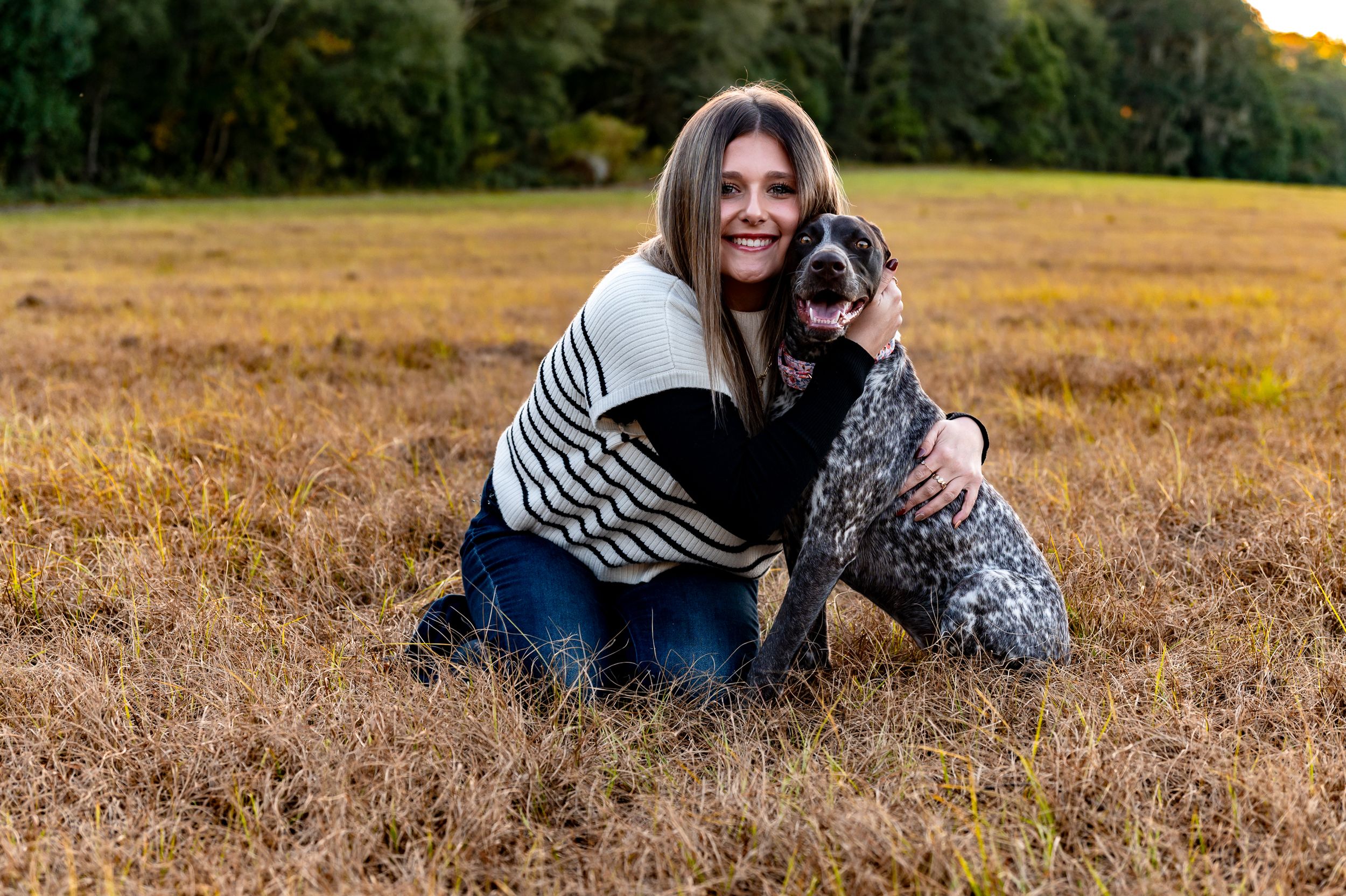 A person in a striped sweater kneels beside their dog in a golden autumn field at sunset.