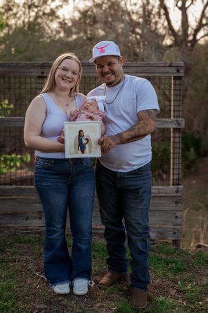 A couple smiles outdoors holding their newborn baby and a pregnancy photo in a wooden fence setting.