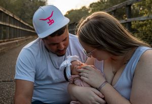 New parents sitting on a bridge, lovingly gazing at their newborn baby during a golden hour outdoor photo session.