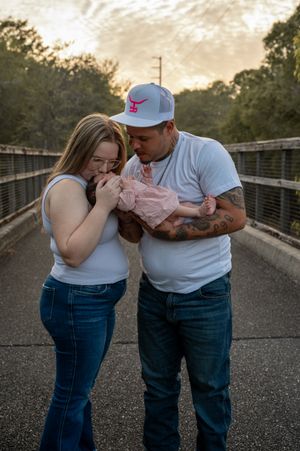 A couple in white tops and jeans admires their newborn baby in pink on a bridge at sunset.