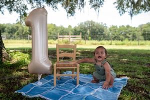 Baby's first birthday celebration outdoors with a large number one balloon and small wooden chair on a blue quilt.