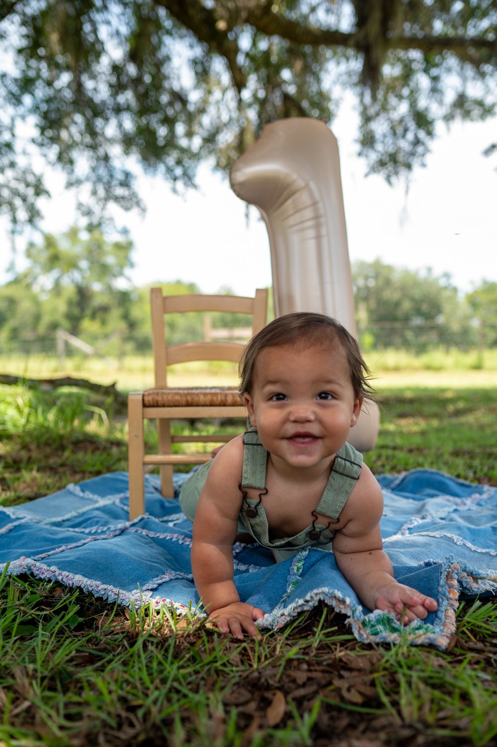 A smiling baby in green overalls lies on a blue blanket outdoors, with a number 1 balloon and wooden chair behind them.