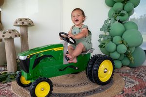 Happy baby sitting on a John Deere ride-on toy tractor at a farm-themed birthday party with green balloon arch.