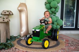 Baby sitting on a John Deere ride-on toy tractor at a first birthday party with green balloon decorations.