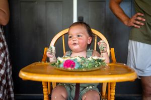 Baby in high chair celebrates first birthday with smashed cake, wearing green outfit, surrounded by family.