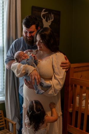 A couple smiles at their newborn baby in a nursery while a young child reaches up toward them.