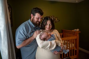 New parents smiling and holding their newborn baby in a nursery with green walls and a wooden crib.