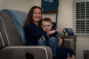 A smiling woman and young boy in navy blue sit together in a gray rocking chair in a cozy room.