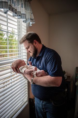 A bearded father tenderly holds his newborn baby near a sunlit window with white blinds.