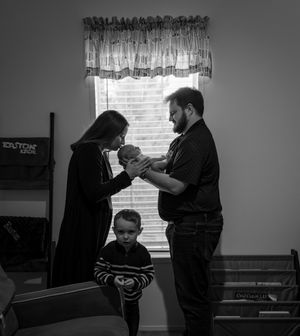 Black and white family photo of parents holding newborn baby while young boy stands between them near window.