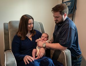 A couple sits together in a chair, lovingly holding their newborn baby in a cozy room.