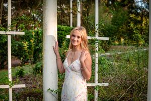 Young blonde woman in floral sundress smiling beside white garden trellis columns surrounded by lush greenery.