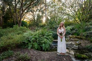 Bride in white lace gown holding bouquet stands in lush garden with ferns and trees at golden hour.