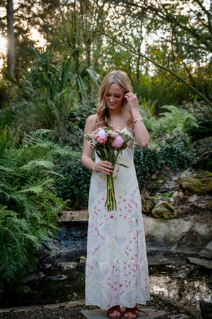 Young blonde woman in floral maxi dress holding pink bouquet, standing in lush garden setting.