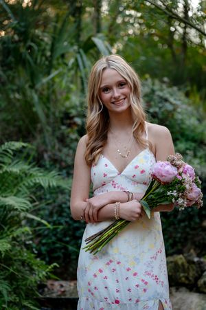 Smiling blonde woman in floral sundress holding pink peony bouquet, standing in lush green garden setting.