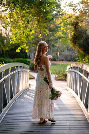 Young woman in floral maxi dress holding pink flowers stands on wooden bridge surrounded by lush green trees.