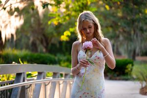 Young blonde woman in floral dress holding a pink peony on a bridge surrounded by lush greenery.