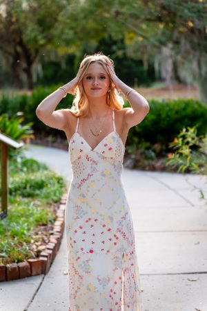 Young woman in floral spaghetti strap maxi dress posing on garden pathway with hands in blonde hair.
