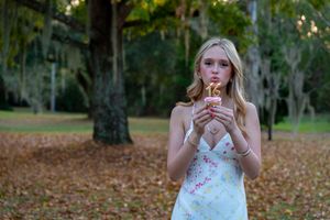 Young blonde girl in floral sundress holding a cupcake outdoors in a park with large trees.