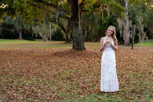 Woman in white floral maxi dress standing among autumn leaves beneath Spanish moss-draped trees in a park.