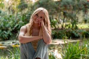 Smiling blonde woman sitting outdoors near a pond, wearing a yellow top and jeans, surrounded by lush green foliage.