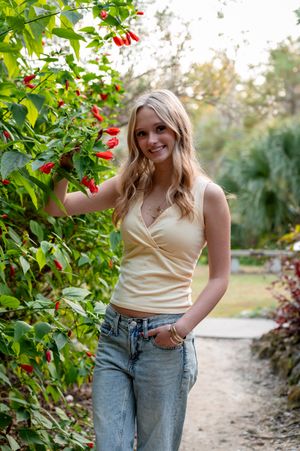 Smiling blonde woman in yellow tank top and jeans posing beside red flowering plants on a garden path.