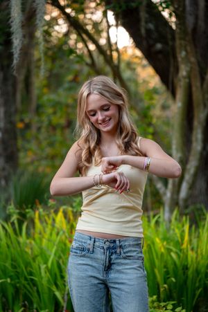 Young blonde woman in yellow tank top and jeans smiling while looking at her bracelets outdoors.