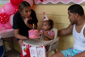 Family celebrating baby's first birthday with pink cake and balloons at a festive outdoor party.