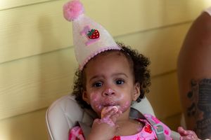 Cute baby girl in pink birthday hat eating cake in high chair, celebrating first birthday with messy frosting face.