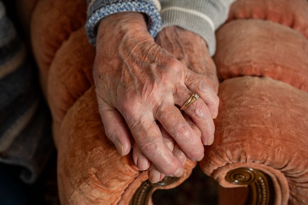 Close-up of elderly hands resting on a pink cushion, showing a wedding ring.