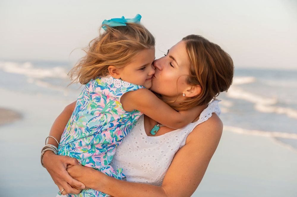 A parent and young child embrace lovingly at the beach during sunset, dressed in summer clothing.