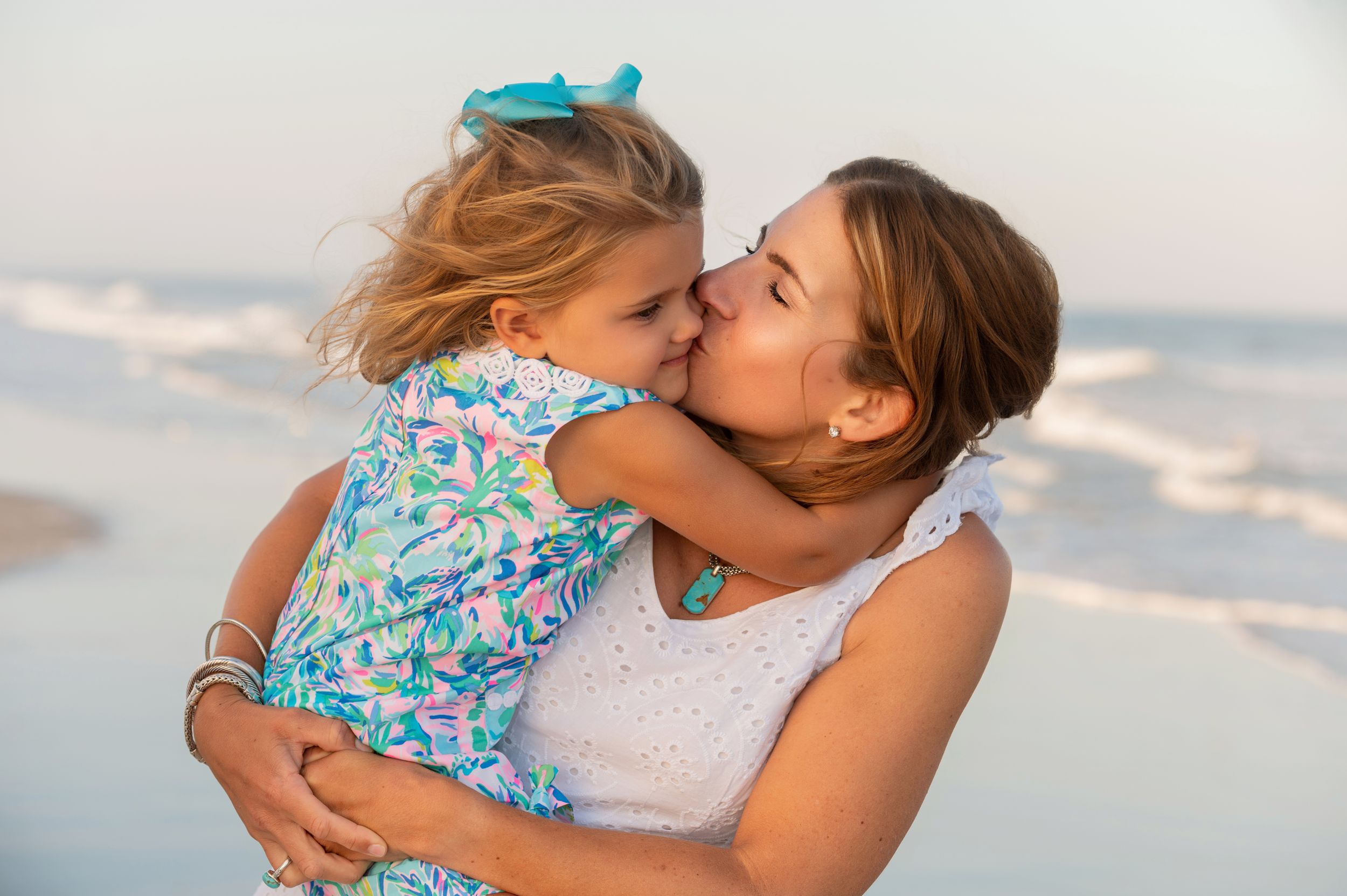 A parent and young child embrace lovingly at the beach during sunset, dressed in summer clothing.