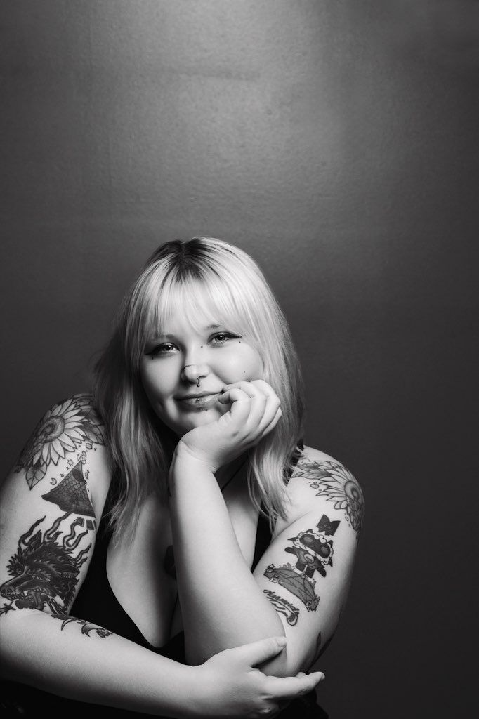 Black and white portrait of someone with blonde hair and tattoos leaning on their hand against dark background.