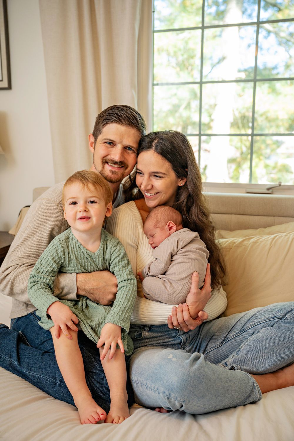 Family portrait on couch by window with toddler and newborn baby in casual setting.