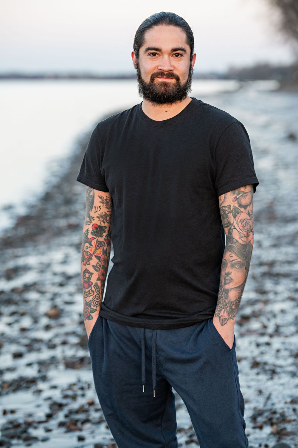 Person with tattoos and beard wearing black t-shirt poses by water at sunset.