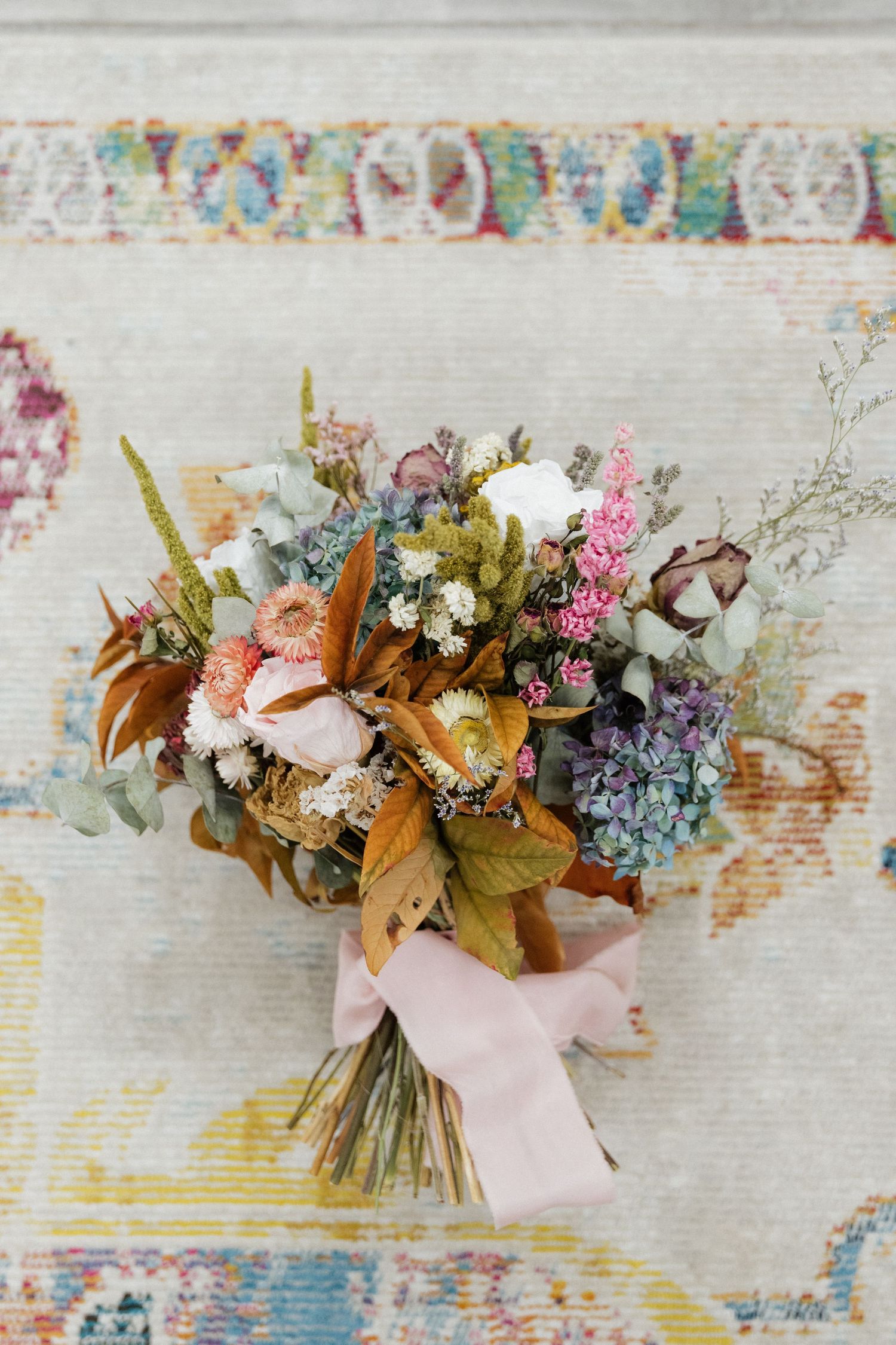 Rustic wedding bouquet with dried flowers in muted autumn colors tied with a pink ribbon on vintage patterned fabric.