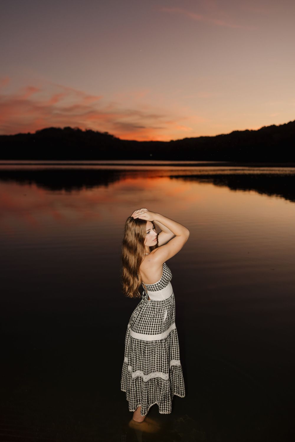 A person in a checkered dress stands silhouetted against a tranquil lake at sunset with mountains in the background.